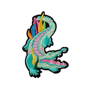 Könnte beinhalten: Ein türkisfarbener Alligator mit rosa und weißen Details und einem Regenbogen aus Federn auf dem Rücken. Der Alligator ist auf einem schwarzen Stoff gestickt.