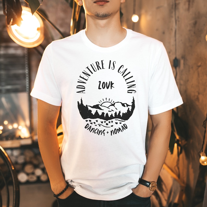 Op de afbeelding: Wit T-shirt met een zwart grafisch ontwerp van een bergketen met een zon en de tekst "Adventure is Calling, Louk, Dancing Nomad".