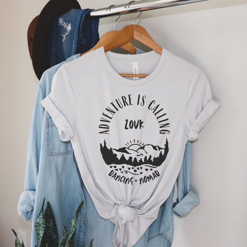 Op de afbeelding: Een wit T-shirt met een zwart grafisch ontwerp met de tekst "Adventure is Calling" met een berglandschap en de woorden "Love, Dancing, Nomad".