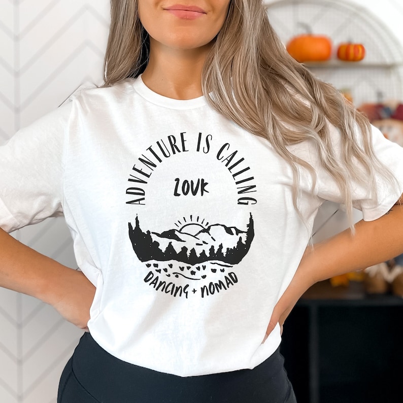 Op de afbeelding: Wit T-shirt met een zwart grafisch ontwerp met de tekst "Adventure is Calling" met een berglandschap en de woorden "Dancing Nomad" eronder.