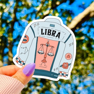May include: A die-cut sticker shaped like a denim jacket in light blue with a peach-colored back panel. The jacket features the word LIBRA in black text above a graphic of a scale. Additional details include small star icons, a rose, and geometric patterns.