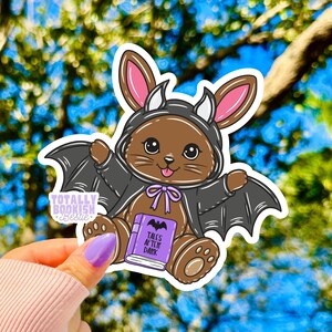 May include: A die-cut sticker featuring a brown cartoon animal wearing a black bat costume with pink-lined ears and wings. The character holds a purple book with the text 'TALES AFTER DARK' and a bat icon. A small logo reads 'TOTALLY BOOKISH Bestie'.
