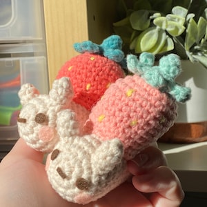 May include: Two crocheted strawberry plushies with embroidered details. One is pink with a blue top and the other is white with brown eyes.