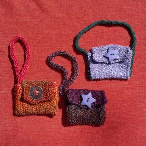 Könnte beinhalten: Drei kleine, handgehäkelte Beutel mit Handschlaufen. Ein Beutel ist orange-grün mit einem silbernen Knopf, einer ist lila mit einem dunkelgrünen Riemen und einem sternförmigen Knopf, und einer ist dunkel mehrfarbig mit einem sternförmigen Knopf.