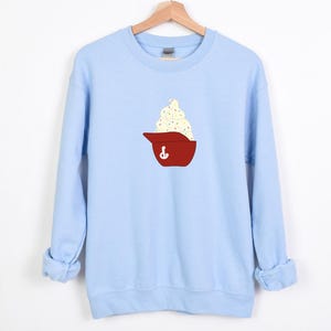 May include: A light blue sweatshirt with a crew neck. The front features a red baseball cap graphic filled with white ice cream and sprinkles. The sweatshirt is made of a soft material.