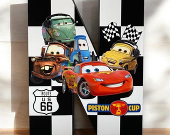 May include: A black and white checkered letter N with a red, yellow, blue, and green car design. The cars are from the Disney Pixar movie Cars. The letter has the text "Route US 66" and "Piston Cup" on it.