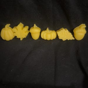 Puede incluir: Seis ceras aromáticas amarillas con formas otoñales: una calabaza, una hoja, una bellota, una calabaza, una hoja y una bellota. Las ceras están dispuestas en fila sobre un fondo negro. Ideal para la decoración temática de otoño.