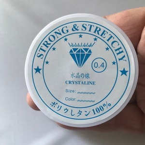 May include: A white round container with the text "STRONG & STRETCHY" in blue lettering. The container also features a blue diamond, the text "水晶の線 CRYSTALINE", and "ボリウレタン100%".