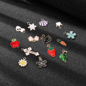 May include: Assortment of 14 gold-tone metal charms with colourful enamel designs. Charms include a star, a crown, a flower, a swan, a cross, a strawberry, a leaf, a bow, an apple, a seashell, a daisy, a flower with a rhinestone, and two abstract shapes.