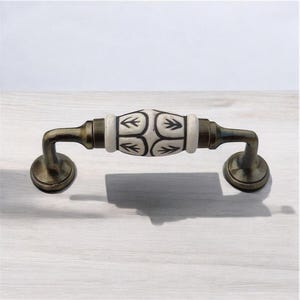 Puede incluir: Un tirador de armario de cerámica blanca con un patrón geométrico negro. El tirador tiene un acabado bronce y está fijado a una superficie de madera.