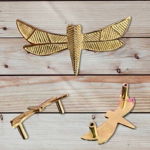 May include: Gold-coloured dragonfly-shaped cabinet pulls. The dragonfly has detailed wing patterns and a textured body. The pulls are shown in different orientations, with mounting hardware visible on some. The background is a light wood grain.