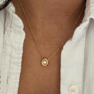 May include: A delicate gold necklace featuring a small, round pearl pendant. The pendant is encased in a gold setting, and the chain is a fine, delicate gold. The necklace is worn against a skin tone and a white button-down shirt.