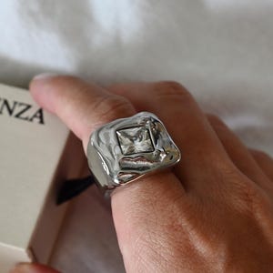 Peut inclure: Une bague argentée avec une texture de surface irrégulière et fondue, ornée d'une pierre centrale transparente de forme carrée. La bague est placée à côté d'une boîte couleur crème avec le texte noir ESSENZA imprimé sur le côté de l'emballage.