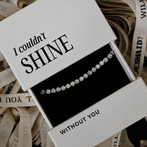 May include: A white gift box with the text "I couldn't SHINE WITHOUT YOU" contains a sparkling tennis bracelet. The box is open, revealing the bracelet against a black interior. Ribbons with text are in the background.