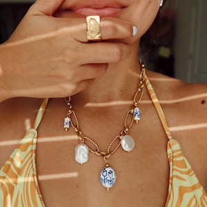 May include: A gold-toned ring with a seashell design and a gold necklace with a chain link design. The necklace features white pearl and blue and white porcelain accents. The necklace has a pendant with a blue floral design.