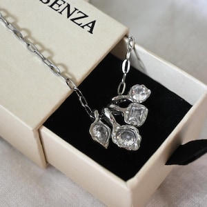 May include: A silver-toned chain necklace featuring four distinct charms, each set with a clear, faceted gemstone. The necklace is presented inside a beige cardboard box with the partial text SENZA printed on the lid, resting on a black insert.