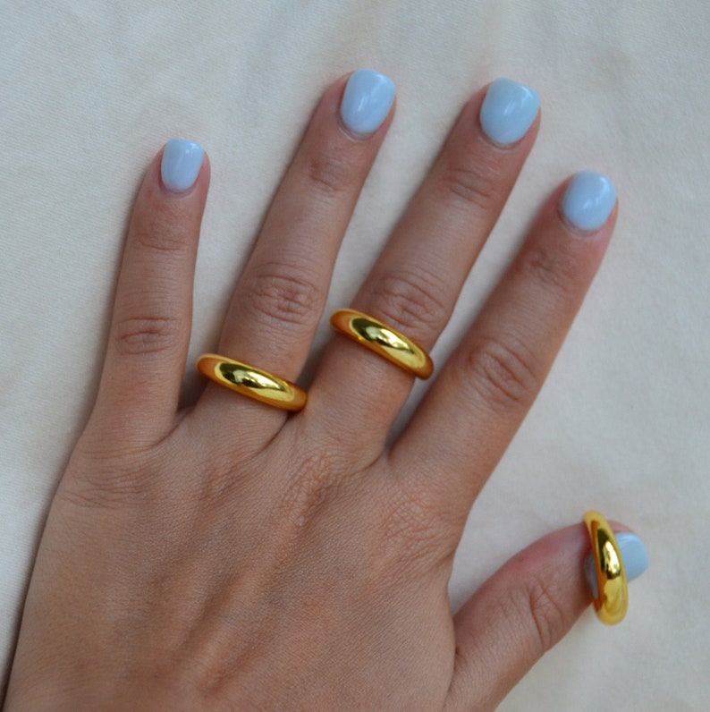 Puede incluir: Tres anillos de color dorado en una mano con esmalte de u&ntilde;as azul claro. Los anillos son de diferentes tama&ntilde;os y se llevan en diferentes dedos.