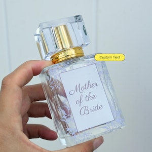 May include: A clear glass bottle of perfume with a gold cap and a white label that says "Mother of the Bride". The label has a floral design around the edges. The bottle is being held by a hand.