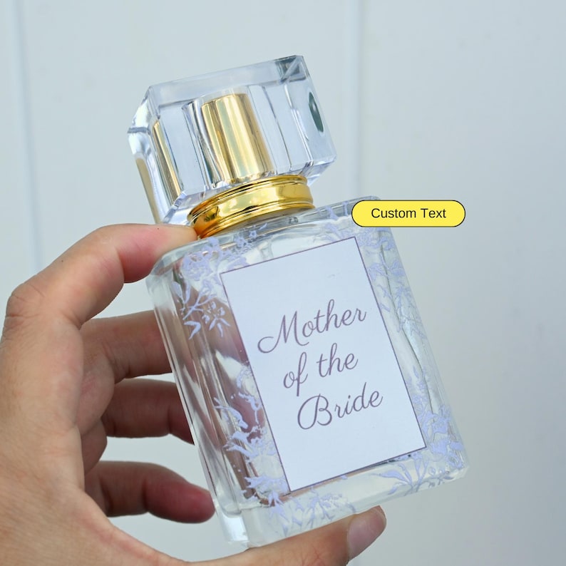 May include: A clear glass bottle of perfume with a gold cap and a white label that says "Mother of the Bride". The label has a floral design around the edges. The bottle is being held by a hand.