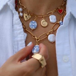May include: Gold-toned necklaces with various charms, including pearls, hearts, and blue and white porcelain pendants. A gold ring is also visible. The jewelry is worn over a white button-down shirt.