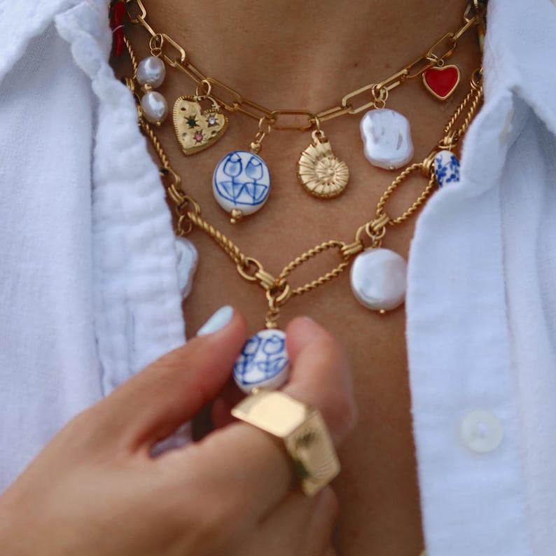 May include: Gold-toned necklaces with various charms, including pearls, hearts, and blue and white porcelain pendants. A gold ring is also visible. The jewelry is worn over a white button-down shirt.