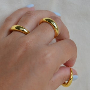 Puede incluir: Tres anillos de color dorado apilados en los dedos de una persona. Los anillos son todos lisos y tienen una forma redondeada.