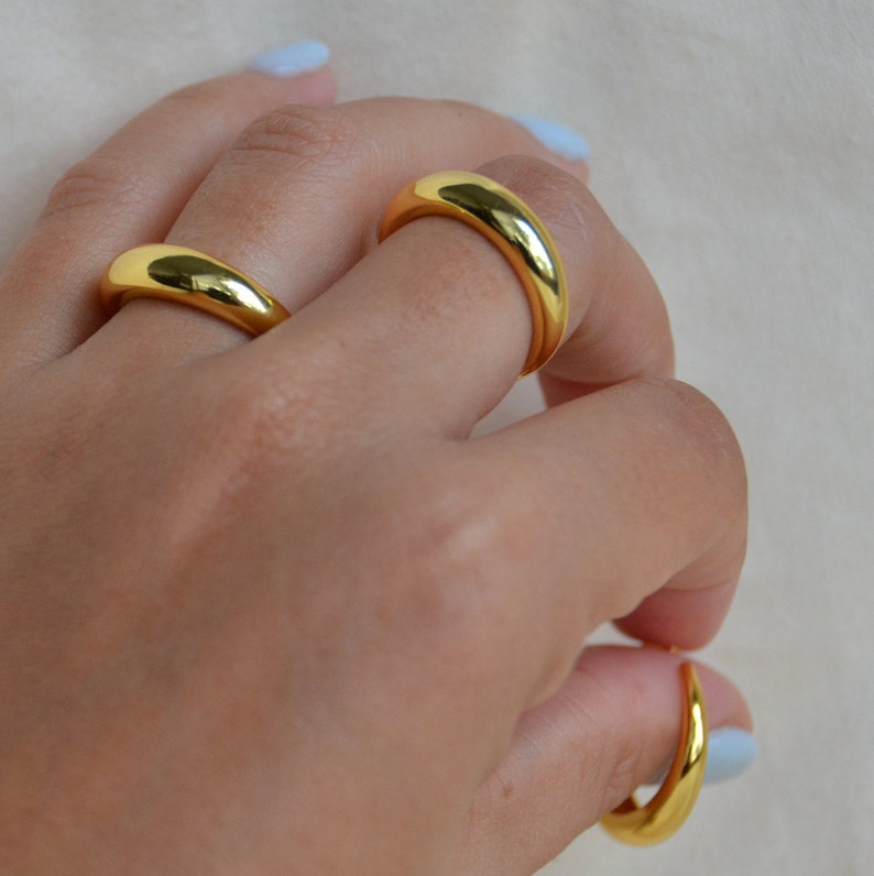 Puede incluir: Tres anillos de color dorado apilados en los dedos de una persona. Los anillos son todos lisos y tienen una forma redondeada.