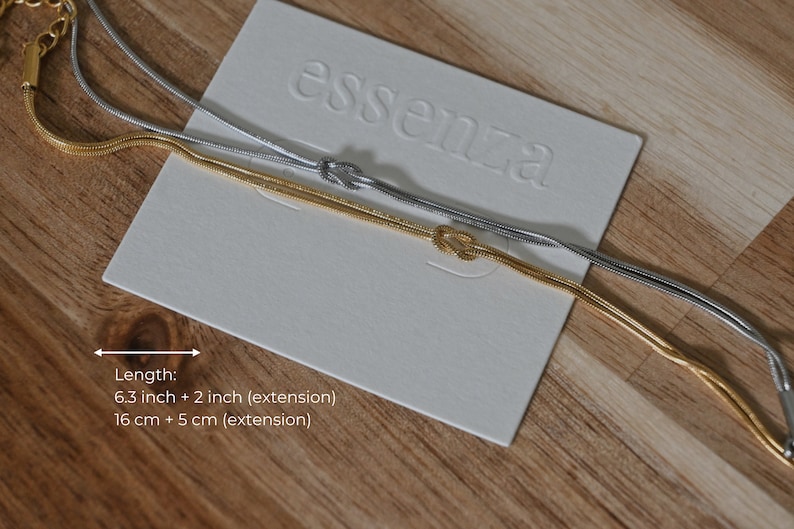 K&ouml;nnte beinhalten: Zwei zarte Kettenhalsb&auml;nder, eines in Gold und eines in Silber, mit einem Knotendesign. Die Halsb&auml;nder liegen auf einer wei&szlig;en Karte mit dem gepr&auml;gten Wort "essenza". Die Karte liegt auf einer Holzoberfl&auml;che. Die L&auml;nge der Halsb&auml;nder betr&auml;gt 6,3 Zoll plus 2 Zoll f&uuml;r die Verl&auml;ngerung, oder 16 cm plus 5 cm f&uuml;r die Verl&auml;ngerung.