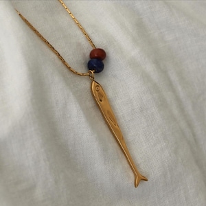 Puede incluir: Un collar de metal en tono dorado con un colgante alargado y fino en forma de pez. La cadena está adornada con dos pequeñas cuentas esféricas en color rojo oscuro y azul oscuro, situadas justo encima del dije metálico de pez.