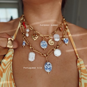 May include: Gold-toned necklace with multiple strands, adorned with pearls, seashell charms, and blue and white ceramic pendants. A gold ring with a seashell design is visible. The text "European summer" and "Portuguese tile" are also visible.
