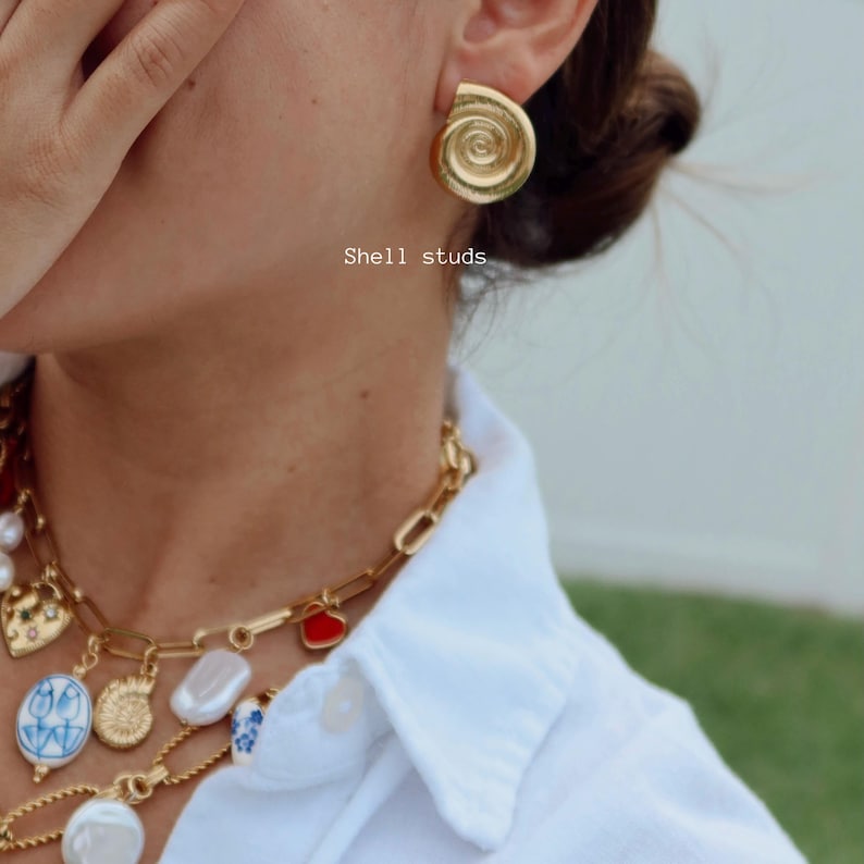 May include: Gold-colored shell stud earrings. The earrings are in the shape of a spiral shell. The image also shows a gold chain necklace with various charms and a white shirt. The text "Shell studs" is visible.