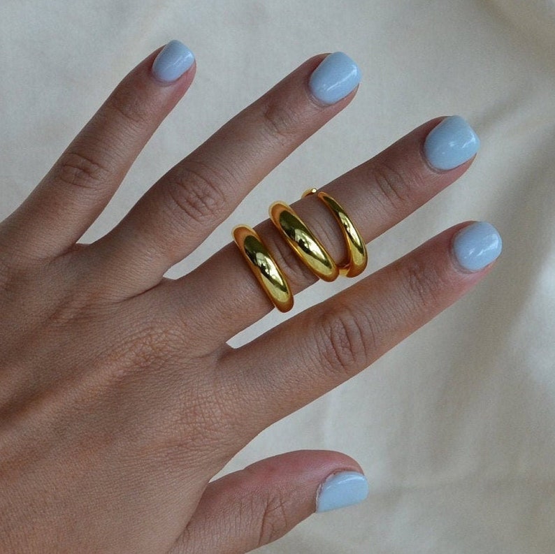 Puede incluir: Tres anillos de color dorado apilados en un dedo. Los anillos son gruesos y tienen una forma redondeada.