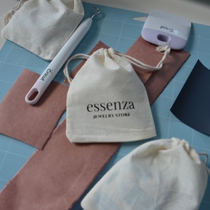 Puede incluir: Una bolsa de tela blanca con cierre de cord&oacute;n. La bolsa tiene el texto "essenza JEWELRY STORE" impreso en ella. La bolsa est&aacute; sobre una estera de corte azul con otros suministros de artesan&iacute;a.