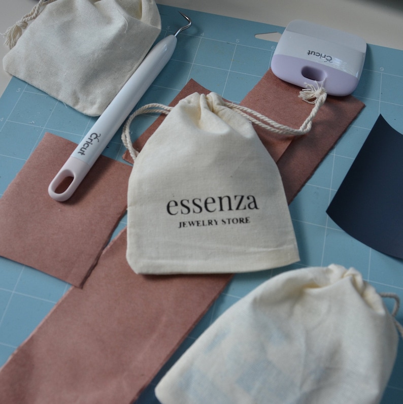 Puede incluir: Una bolsa de tela blanca con cierre de cord&oacute;n. La bolsa tiene el texto "essenza JEWELRY STORE" impreso en ella. La bolsa est&aacute; sobre una estera de corte azul con otros suministros de artesan&iacute;a.