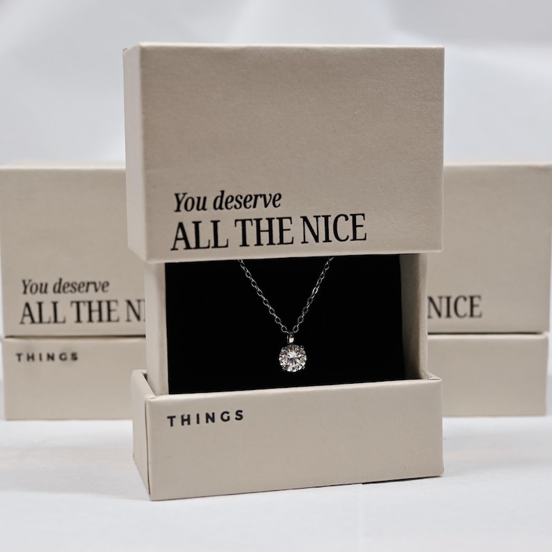 Pu&ograve; includere: Una scatola bianca con il testo "You deserve ALL THE NICE THINGS" sulla parte superiore e una collana d'argento con un ciondolo rotondo all'interno. La scatola &egrave; su una superficie bianca.