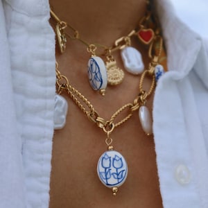 May include: Gold-toned chain necklaces with various charms, including a red heart, a sunburst, and white baroque pearls. The necklaces also feature blue and white ceramic oval pendants with floral designs.