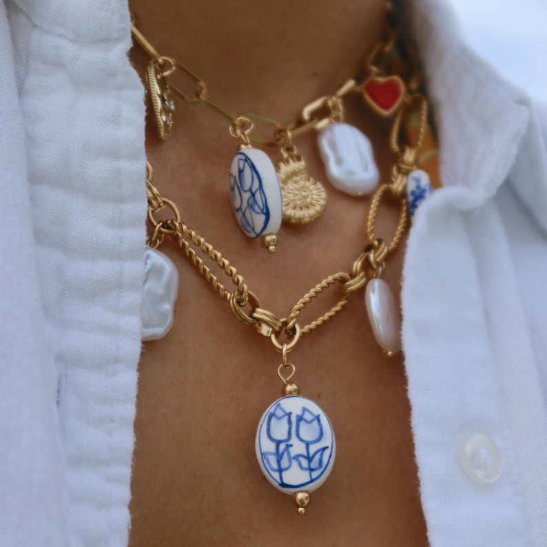 May include: Gold-toned chain necklaces with various charms, including a red heart, a sunburst, and white baroque pearls. The necklaces also feature blue and white ceramic oval pendants with floral designs.