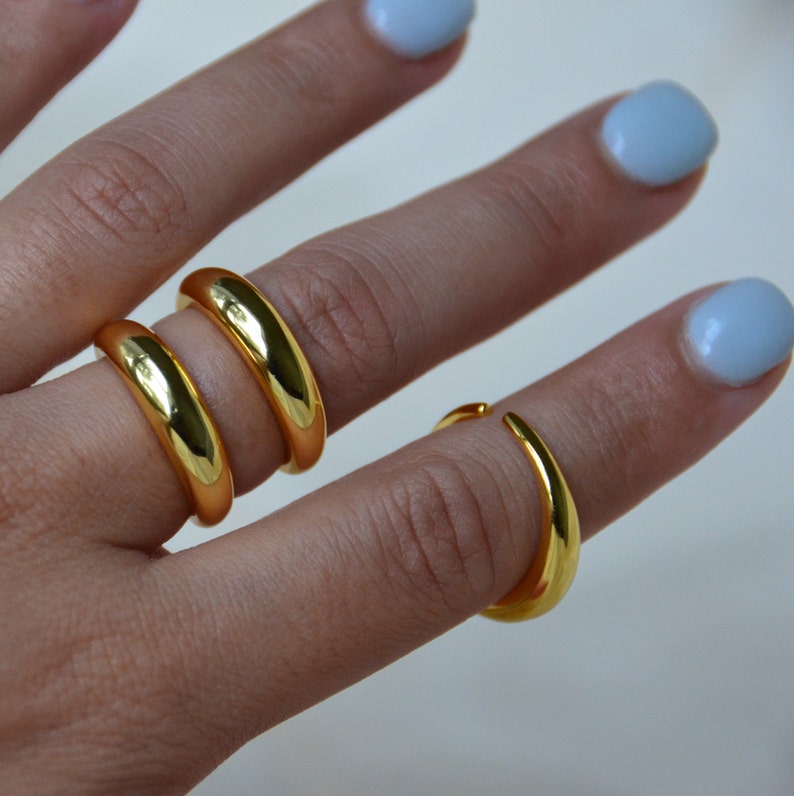 Puede incluir: Tres anillos de oro apilados en un dedo. Los anillos son todos lisos y tienen una forma redondeada.