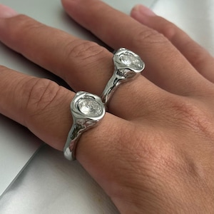 May include: Two silver rings with a unique, organic design. Each ring features a clear, round gemstone set in a textured, molten-like setting. The rings are displayed on a hand, showcasing their reflective qualities and intricate details.