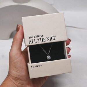 Pu&ograve; includere: Una scatola bianca con il testo "You deserve ALL THE NICE THINGS" sulla parte superiore. La scatola contiene una collana d'argento con un ciondolo rotondo di pietra trasparente.