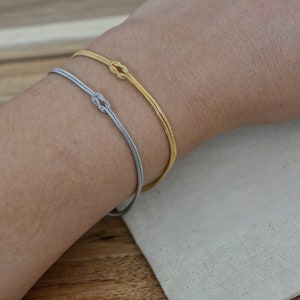 K&ouml;nnte beinhalten: Zwei Armb&auml;nder am Handgelenk einer Person. Das obere Armband ist golden und das untere Armband ist silber.