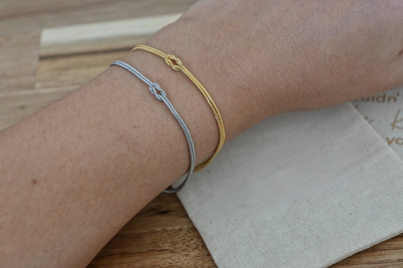 K&ouml;nnte beinhalten: Zwei Armb&auml;nder am Handgelenk einer Person. Das obere Armband ist golden und das untere Armband ist silber.