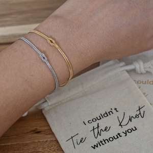 Op de afbeelding: Twee armbanden met knoopdetails, een zilveren en een gouden, om een pols. De armbanden worden getoond op een witte stoffen tas met de tekst "Ik kon de knoop niet zonder jou strikken".