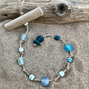May include: A handcrafted necklace made with sea glass beads in shades of blue and white, strung on a natural jute cord. The necklace is displayed on a sandy surface with a piece of driftwood, creating a beachy aesthetic.