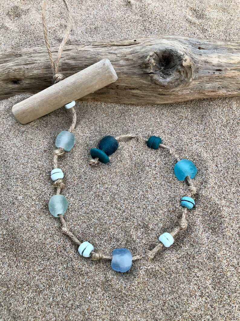 May include: A handcrafted necklace made with sea glass beads in shades of blue and white, strung on a natural jute cord. The necklace is displayed on a sandy surface with a piece of driftwood, creating a beachy aesthetic.