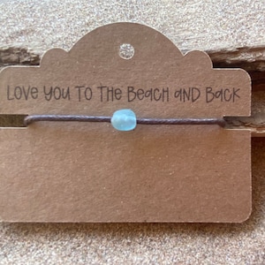 Pode incluir: Uma pulseira de cord&atilde;o castanho com uma conta azul clara &eacute; exibida em um cart&atilde;o castanho. O cart&atilde;o tem a frase "Love you To The Beach and Back" impressa em preto. O fundo &eacute; uma superf&iacute;cie texturizada castanho claro.