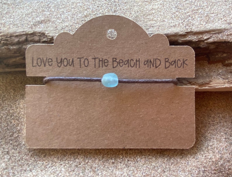 Pode incluir: Uma pulseira de cord&atilde;o castanho com uma conta azul clara &eacute; exibida em um cart&atilde;o castanho. O cart&atilde;o tem a frase "Love you To The Beach and Back" impressa em preto. O fundo &eacute; uma superf&iacute;cie texturizada castanho claro.