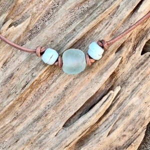 May include: A simple necklace with a light blue sea glass bead and two white beads on a brown leather cord.