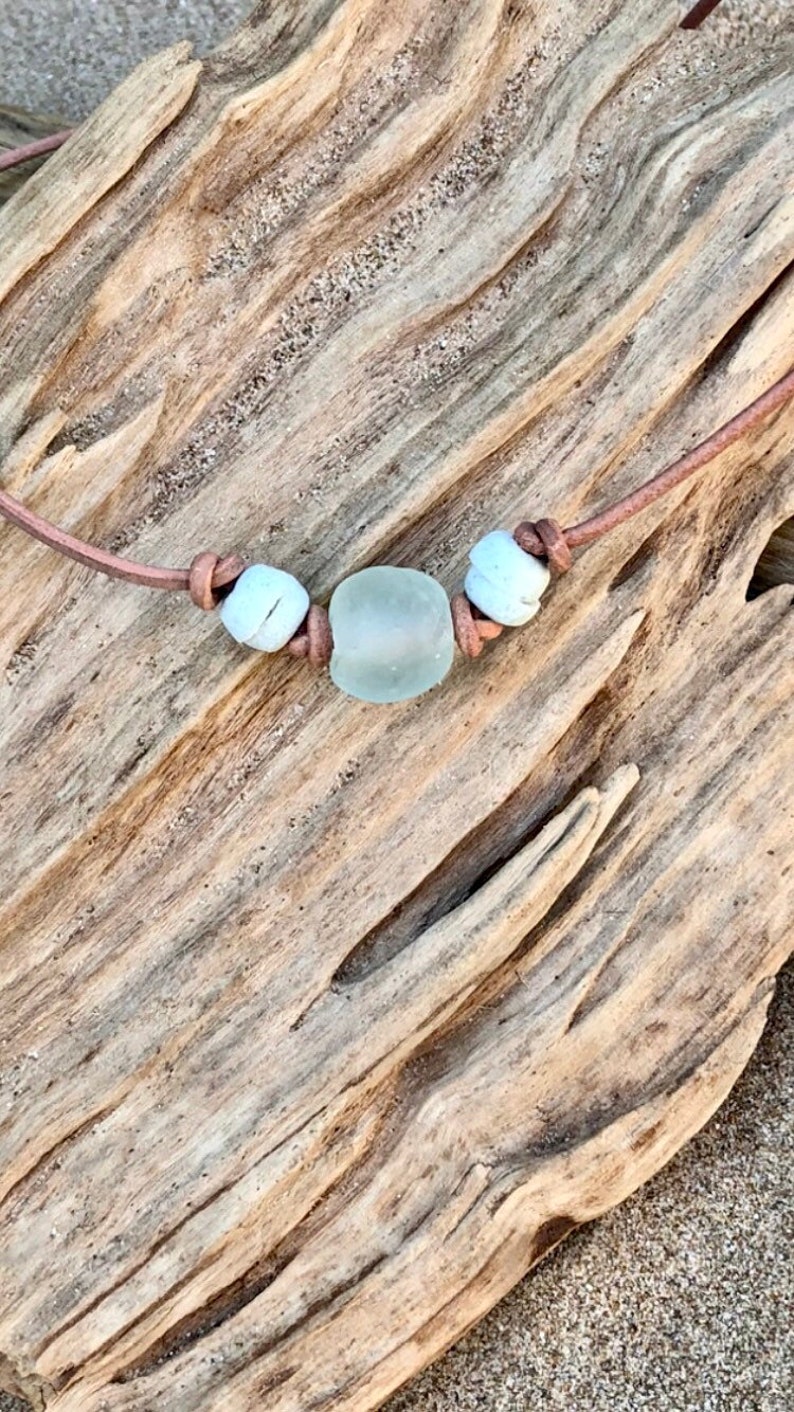 May include: A simple necklace with a light blue sea glass bead and two white beads on a brown leather cord.