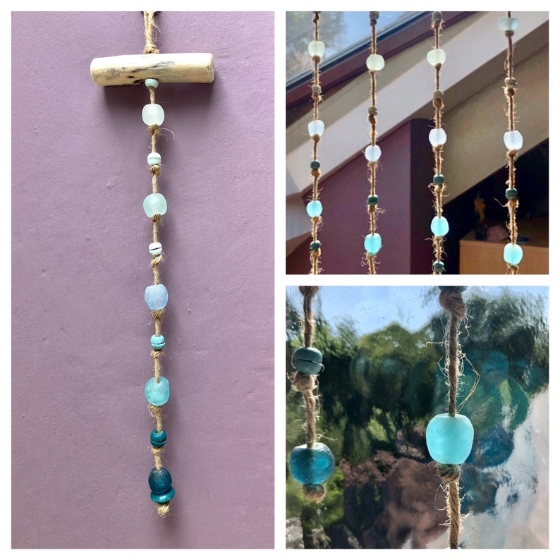 May include: A decorative hanging ornament featuring a natural wood piece and a jute string. The string is adorned with various sea glass beads in shades of blue, green, and white. The beads vary in size and shape, creating a unique coastal aesthetic.